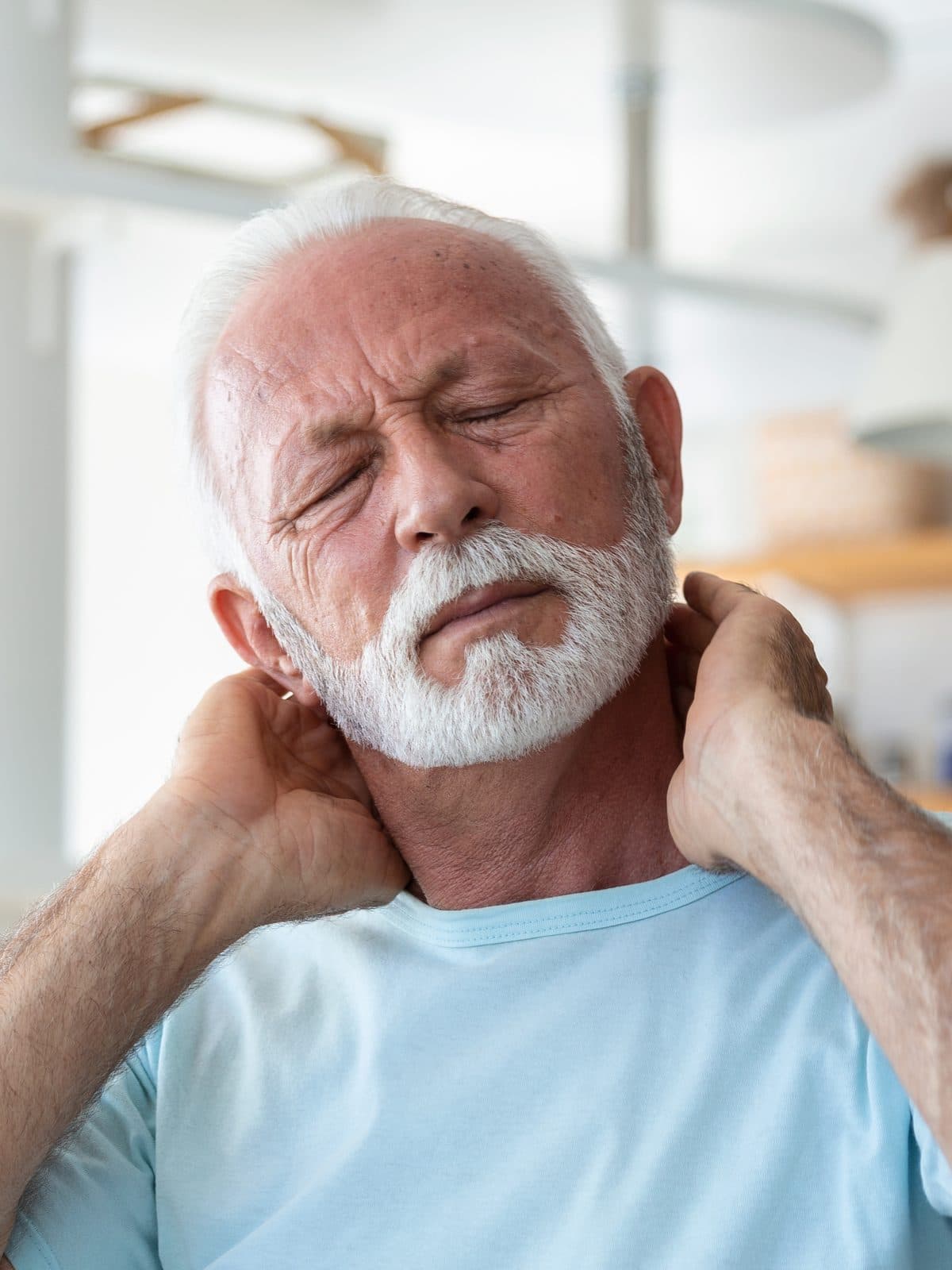 A senior man wincing and holding the back of his neck with both hands.