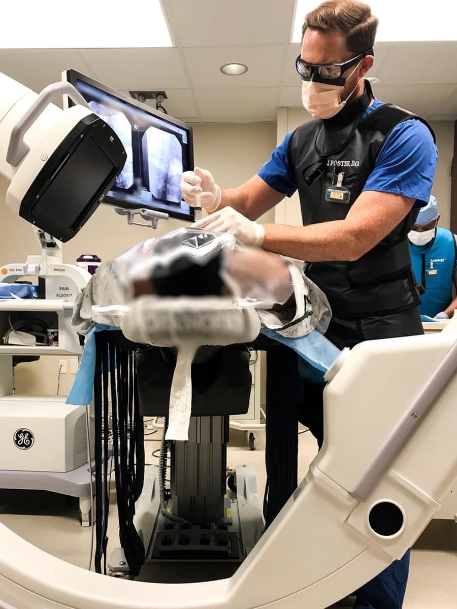 Dr. Foster at a C-arm fluoroscopy setup, working alongside a radiology technician.