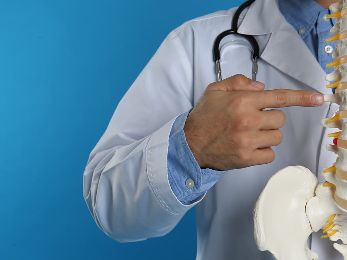 A physician in a white coat with a stethoscope, pointing to a vertebra on a spine model.